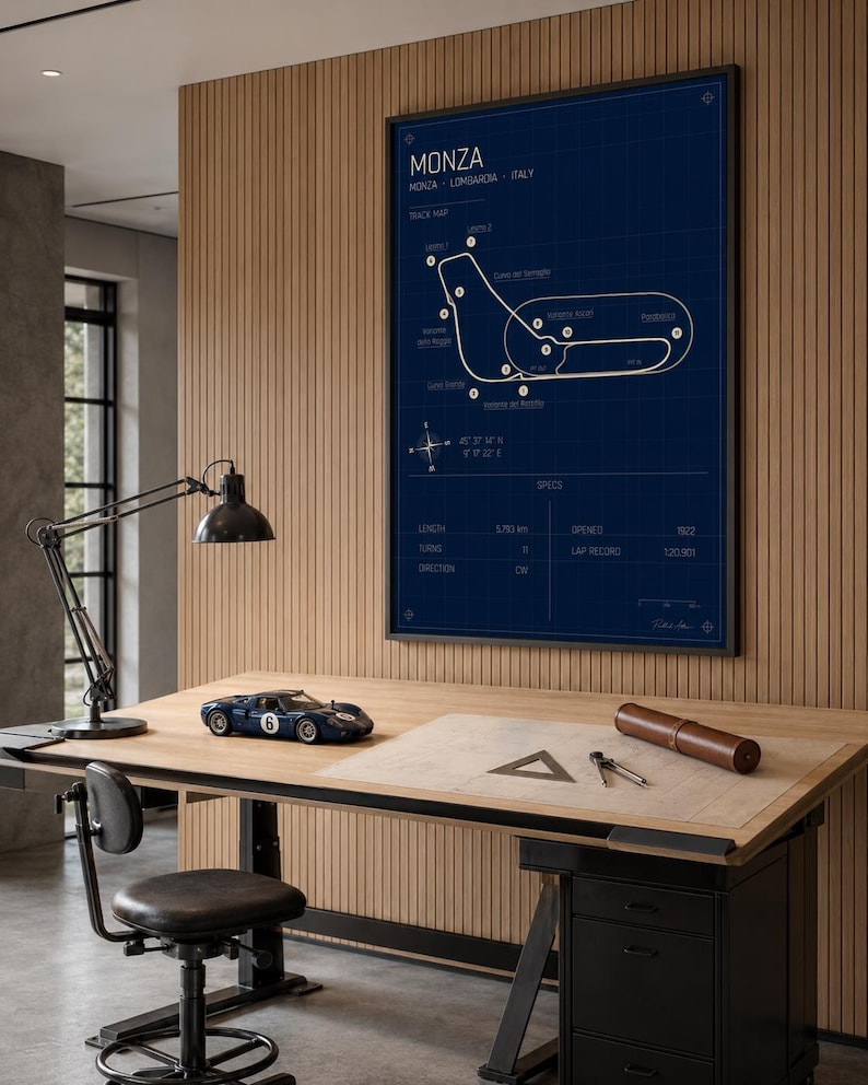Op de afbeelding: Een ingelijste blauwdruk-stijl poster van het Monza circuit in Itali&euml;, met een donkerblauwe achtergrond en witte lijnen. Een klein blauw racewagenmodel staat op een houten bureau, samen met tekengereedschap en een bureaulamp.