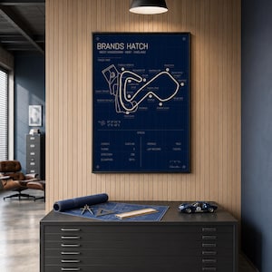 May include: A framed Brands Hatch race track map in navy blue, with white lines and text. A rolled-up blueprint, drafting tools, and a blue toy race car sit on a dark gray cabinet with drawers. The text on the map includes "BRANDS HATCH" and "WEST KINGSDOWN - KENT - ENGLAND."