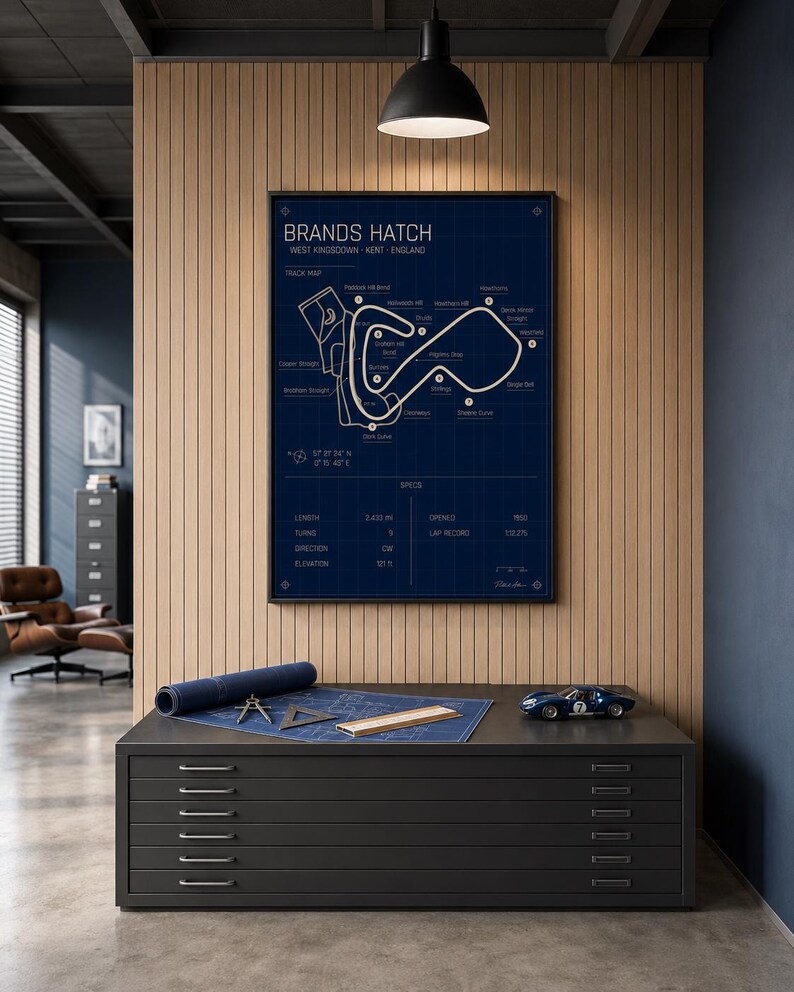 May include: A framed Brands Hatch race track map in navy blue, with white lines and text. A rolled-up blueprint, drafting tools, and a blue toy race car sit on a dark gray cabinet with drawers. The text on the map includes "BRANDS HATCH" and "WEST KINGSDOWN - KENT - ENGLAND."