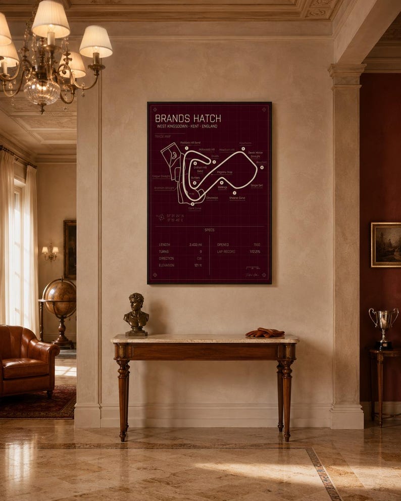 May include: A framed burgundy print of the Brands Hatch race track in West Kingsdown, Kent, England. The track map is white, with text at the top and bottom. The print hangs on a beige wall above a wooden table.