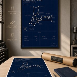 K&ouml;nnte beinhalten: Gerahmter Cadwell Park Streckenplan-Druck in Marineblau mit wei&szlig;em Text und Rasterhintergrund. Der Druck enth&auml;lt Streckendetails und Spezifikationen. Eine gerollte Druck, ein Notizbuch und Zeichenwerkzeuge liegen auf dem Tisch.