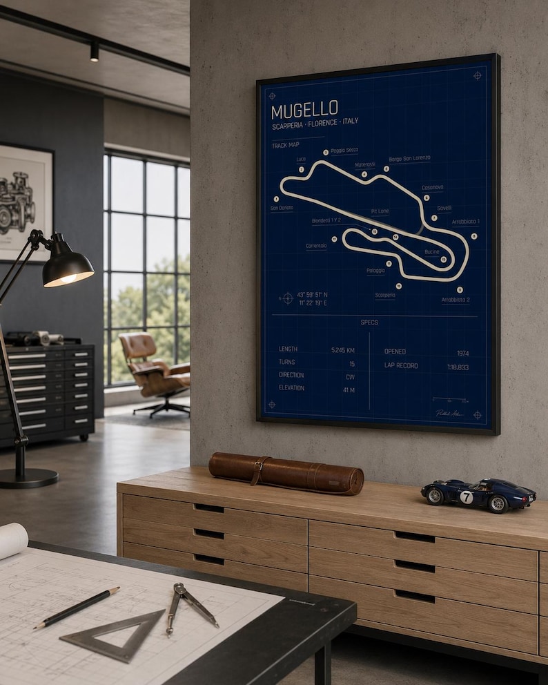 May include: A framed blueprint-style print of the Mugello race track in Italy, with the track layout in white against a dark blue background. The print includes track specifications and is displayed above a wooden cabinet. A small blue race car model sits on the cabinet.