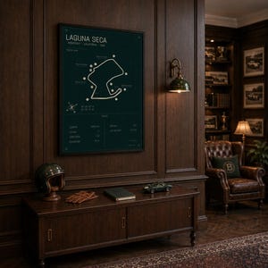 May include: A dark green framed poster of the Laguna Seca racetrack in Monterey, California, USA, hangs on a dark wood paneled wall. A vintage helmet, gloves, and a model car sit on a wooden cabinet below.