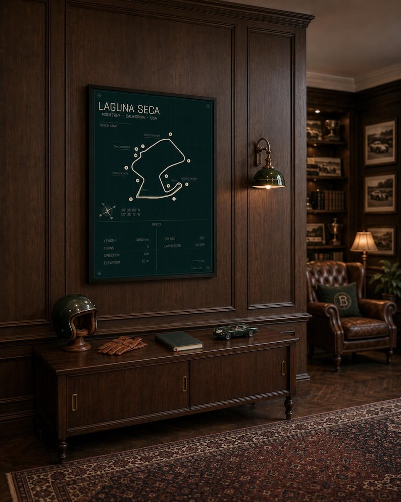 May include: A dark green framed poster of the Laguna Seca racetrack in Monterey, California, USA, hangs on a dark wood paneled wall. A vintage helmet, gloves, and a model car sit on a wooden cabinet below.