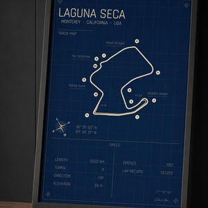 May include: Framed Laguna Seca race track map in a black frame. The map is white on a dark blue background, with text including "Laguna Seca" and track details. The frame is on a wooden surface.