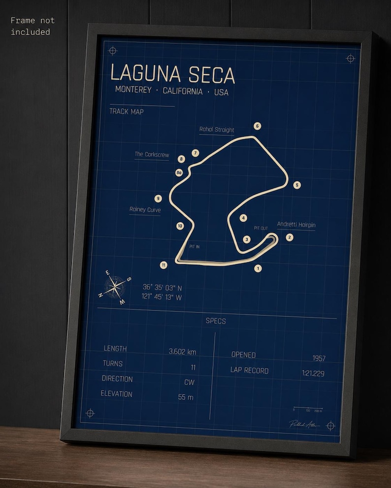 May include: Framed Laguna Seca race track map in a black frame. The map is white on a dark blue background, with text including "Laguna Seca" and track details. The frame is on a wooden surface.