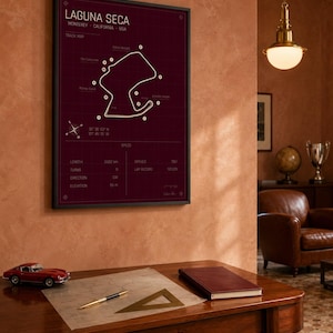 May include: A framed print of the Laguna Seca racetrack in Monterey, California, USA. The track map is white on a dark red background. A red toy car, a pen, and a notebook sit on a wooden desk.