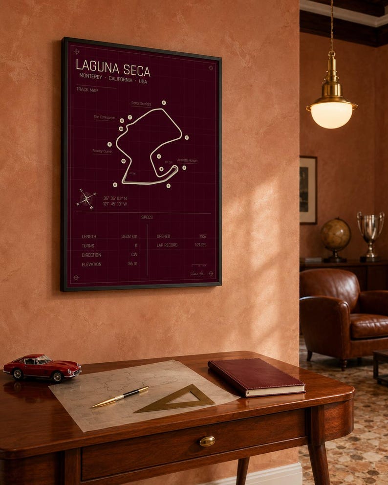 May include: A framed print of the Laguna Seca racetrack in Monterey, California, USA. The track map is white on a dark red background. A red toy car, a pen, and a notebook sit on a wooden desk.