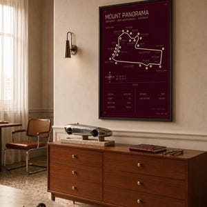 May include: A framed Mount Panorama race track map in burgundy with white detailing hangs above a wooden dresser. A silver model race car sits on the dresser, along with a leather-bound book and a small red race car model.