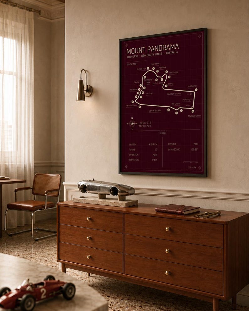 May include: A framed Mount Panorama race track map in burgundy with white detailing hangs above a wooden dresser. A silver model race car sits on the dresser, along with a leather-bound book and a small red race car model.