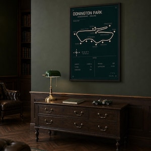 Op de afbeelding: Een ingelijste, donkergroene print van het Donington Park-circuit in Leicestershire, Engeland, hangt boven een houten dressoir. De print bevat een circuitkaart en specificaties. Een groene bureaulamp en een speelgoedraceauto staan op het dressoir.