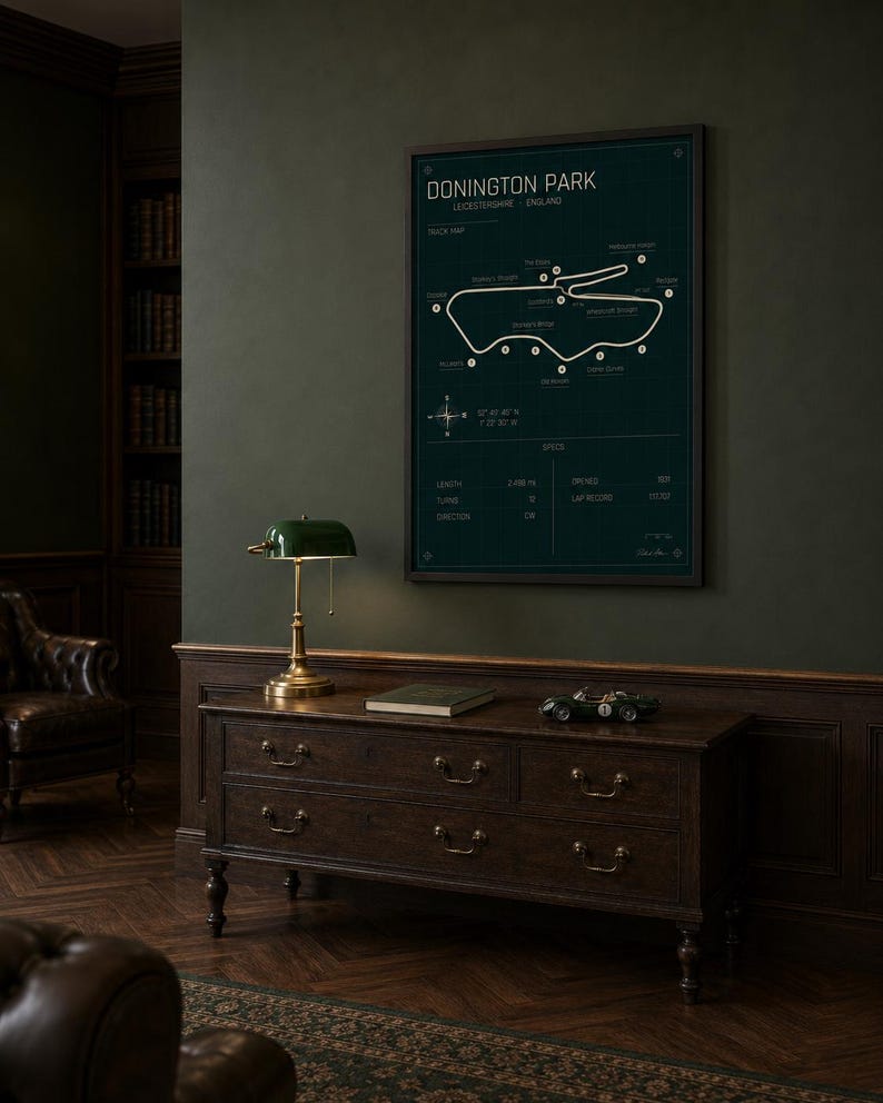 Op de afbeelding: Een ingelijste, donkergroene print van het Donington Park-circuit in Leicestershire, Engeland, hangt boven een houten dressoir. De print bevat een circuitkaart en specificaties. Een groene bureaulamp en een speelgoedraceauto staan op het dressoir.