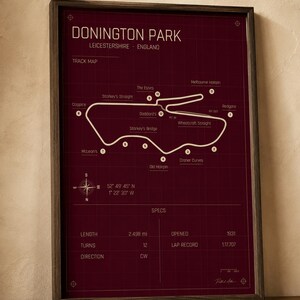 Op de afbeelding: Een ingelijste print van het Donington Park circuit in Leicestershire, Engeland. De kaart is wit op een bordeauxrode achtergrond, met details zoals lengte, bochten en ronderecord. De lijst is donkerbruin.