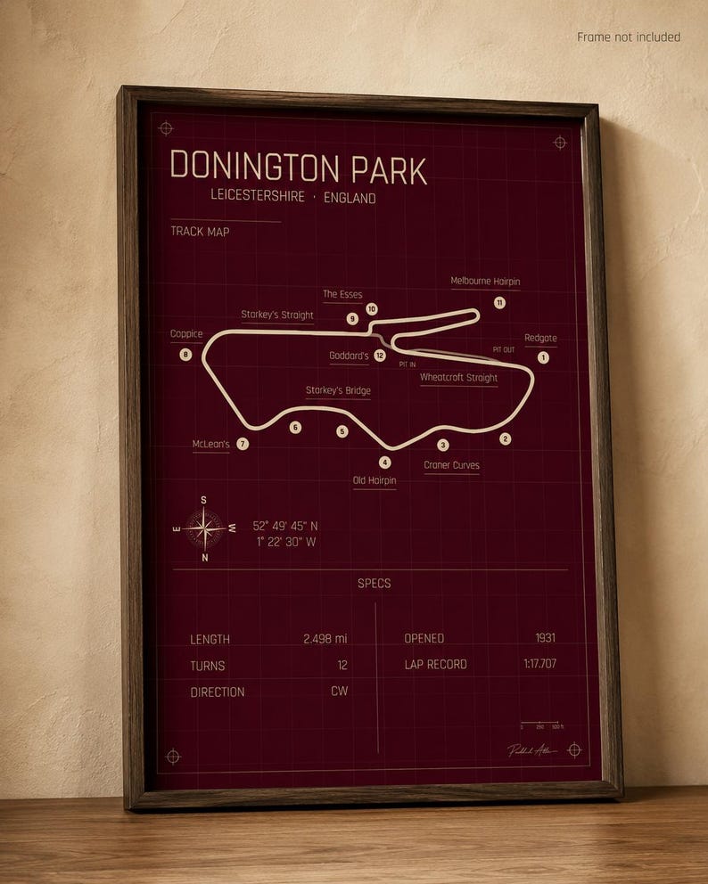 Op de afbeelding: Een ingelijste print van het Donington Park circuit in Leicestershire, Engeland. De kaart is wit op een bordeauxrode achtergrond, met details zoals lengte, bochten en ronderecord. De lijst is donkerbruin.