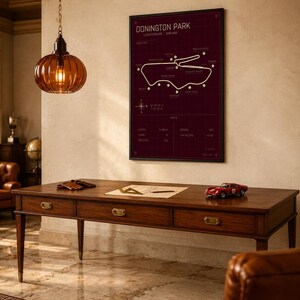 Op de afbeelding: Een ingelijste Donington Park-circuitkaart in een kamer met een houten bureau, een rode speelgoedauto en een hangende amberkleurige glazen lamp. De kaart is wit op een donkerrode achtergrond. Het bureau heeft drie laden.