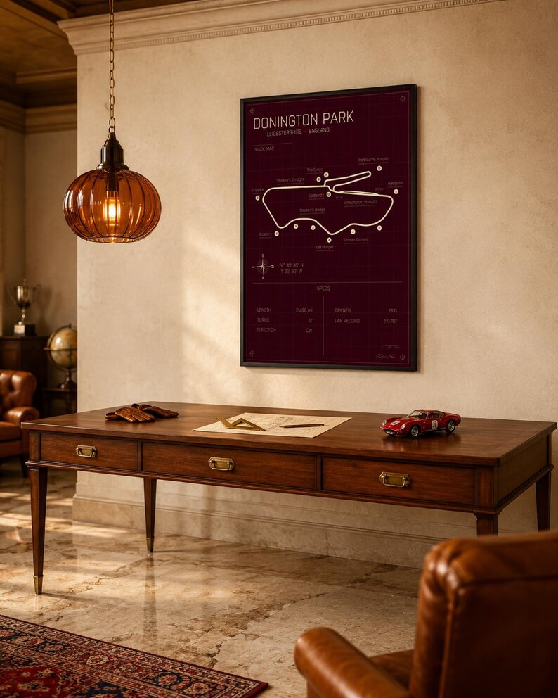 Op de afbeelding: Een ingelijste Donington Park-circuitkaart in een kamer met een houten bureau, een rode speelgoedauto en een hangende amberkleurige glazen lamp. De kaart is wit op een donkerrode achtergrond. Het bureau heeft drie laden.