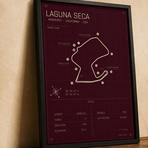 May include: Framed print of the Laguna Seca racetrack in Monterey, California, USA. The track map is white on a burgundy background, with details including length (3.602 km), turns (11), and elevation (55 m). The frame is black.