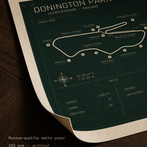 Op de afbeelding: Een donkergroene print van het Donington Park circuit in Leicestershire, Engeland. De circuitkaart bevat gelabelde bochten en een kompasroos. De print bevat ook specificaties zoals lengte en bochten. De tekst "Museum-quality matte paper" is zichtbaar.