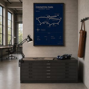 Op de afbeelding: Een ingelijste marineblauwe print van het Donington Park circuit in Leicestershire, Engeland. De circuitkaart is wit op een marineblauwe achtergrond. Een zwarte metalen kast met lades en een blauw modelraceauto staan op de voorgrond.