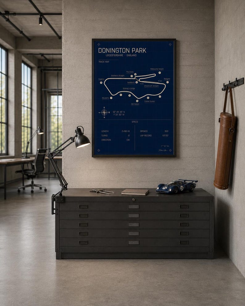 Op de afbeelding: Een ingelijste marineblauwe print van het Donington Park circuit in Leicestershire, Engeland. De circuitkaart is wit op een marineblauwe achtergrond. Een zwarte metalen kast met lades en een blauw modelraceauto staan op de voorgrond.