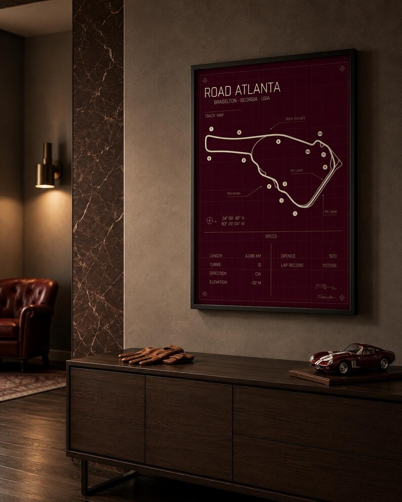 May include: Framed print of the Road Atlanta racetrack in Braselton, Georgia, USA. The track map is white on a burgundy background, with specifications listed below. A model race car and driving gloves sit on a dark wood cabinet.