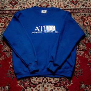 May include: A royal blue sweatshirt with the text "ATI ADVANCED TECHCOM, INC." in white. The sweatshirt has a crew neck and long sleeves. The brand label is visible on the upper back. The sweatshirt is displayed on a patterned rug.
