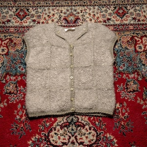 May include: A light gray, short-sleeved cardigan sweater with a button-down front. The sweater has a textured knit pattern and two small pockets. The buttons are a matching light gray color. The sweater is displayed on a patterned red rug.