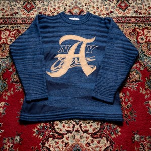 Peut inclure: Un pull en tricot bleu à manches longues avec une texture côtelée et une grande application beige sur la poitrine. L'application affiche la lettre A avec les lettres N et Y de chaque côté et le mot Avirex écrit en cursive en dessous.