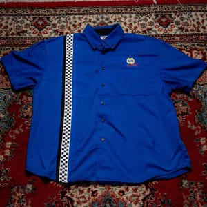Peut inclure: Une chemise à manches courtes bleu à boutons, dotée d'une bande de course verticale à damier noir et blanc sur le côté gauche. La chemise comprend un col, une poche avant et un logo NAPA Racing jaune et bleu brodé sur la poitrine.