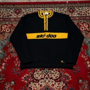 Peut inclure: Un pull noir à manches longues avec une bande horizontale jaune sur la poitrine portant l'inscription ski-doo en noir. Le pull présente un col jaune à rayures noires et une patte de boutonnage partielle avec quatre boutons.