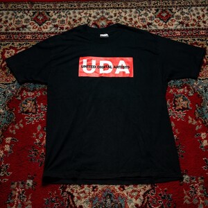 Peut inclure: Un t-shirt noir à manches courtes avec un graphique rectangulaire rouge sur la poitrine. À l'intérieur du rectangle rouge, les lettres blanches UDA sont imprimées en gras, avec le texte UNITED DIGITAL ARTISTS en petites lettres noires.