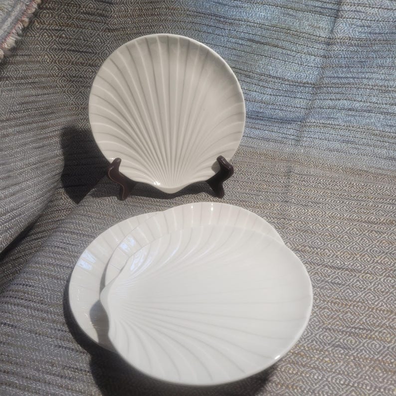 May include: A set of three off-white ceramic plates shaped like scallop shells. One plate is displayed upright on a dark brown stand, while two identical plates are stacked horizontally in front, featuring embossed radial ridges.