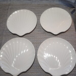 May include: A set of four white ceramic plates, each shaped like a scallop shell. The plates feature a glossy finish and raised, radiating ridges that extend from the base to the scalloped edges, creating a distinct shell-like texture.