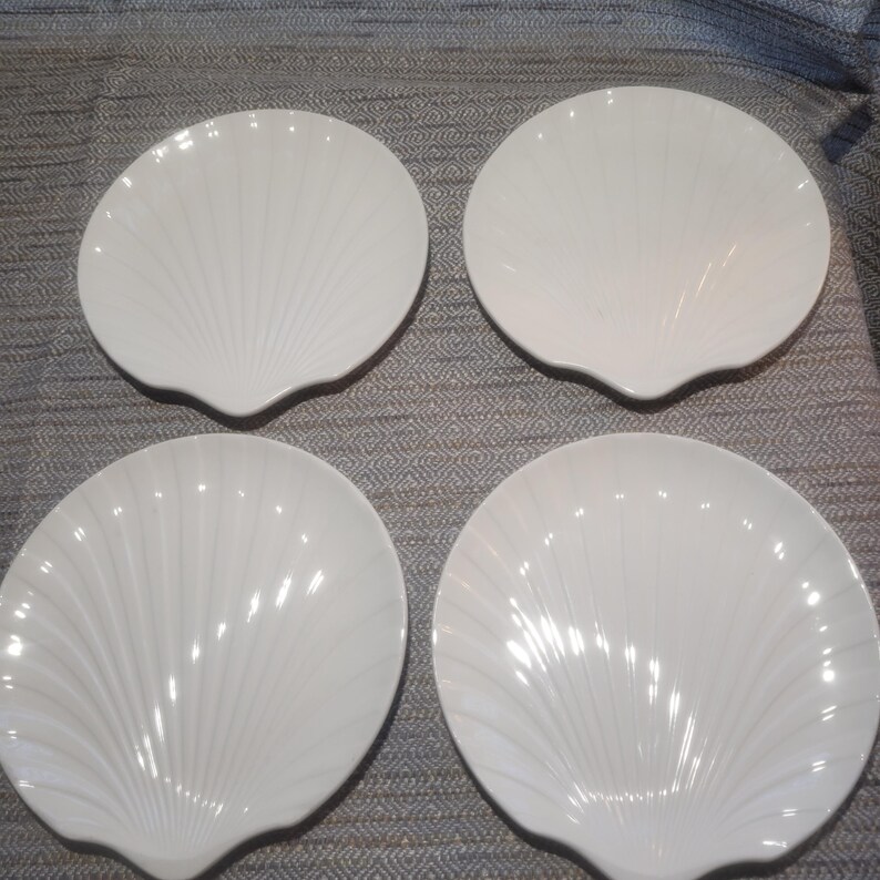 May include: A set of four white ceramic plates, each shaped like a scallop shell. The plates feature a glossy finish and raised, radiating ridges that extend from the base to the scalloped edges, creating a distinct shell-like texture.