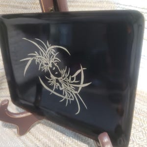 May include: A rectangular black tray with a glossy finish featuring a centered, light-colored, intricate line drawing of stylized grass or floral elements. The tray has rounded corners and a smooth, reflective surface, presented on a stand.
