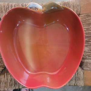 Könnte beinhalten: Eine apfelförmige Keramikschale mit glänzend roter Oberfläche, die in eine hellere gelb-orange Mitte übergeht. Die Schale ist mit einem kleinen, erhabenen grünen Blatt und einem weißen Stiel am oberen Rand versehen, was die Fruchtform betont.
