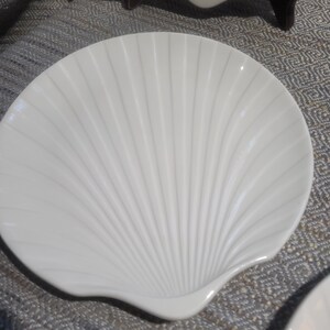 May include: A white ceramic dish shaped like a scallop shell. The surface features a series of raised, radiating ridges that extend from the base to the scalloped edge, creating a textured, fan-like appearance with a smooth, glossy finish.