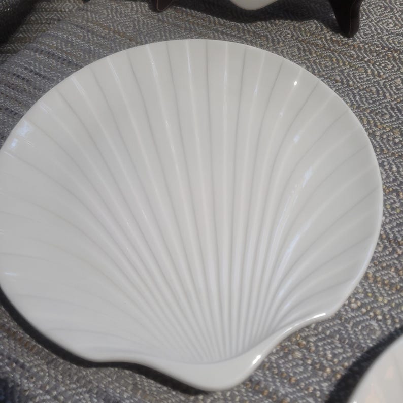 May include: A white ceramic dish shaped like a scallop shell. The surface features a series of raised, radiating ridges that extend from the base to the scalloped edge, creating a textured, fan-like appearance with a smooth, glossy finish.