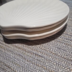May include: A stack of three off-white ceramic plates, each shaped like a seashell with a scalloped edge and radiating embossed lines that mimic the natural texture of a shell. The plates are smooth, glossy, and stacked neatly on top of each other.