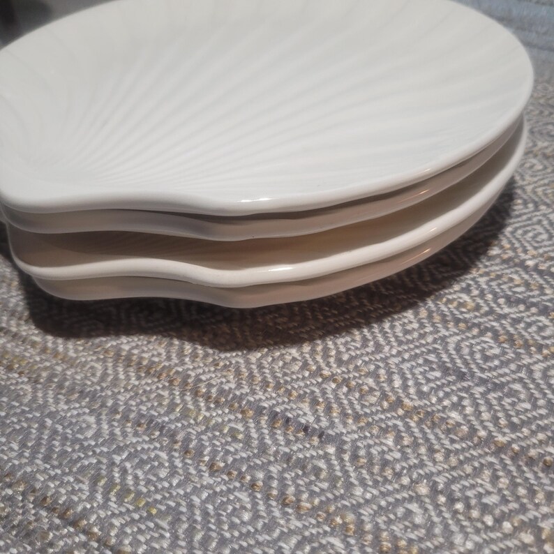 May include: A stack of three off-white ceramic plates, each shaped like a seashell with a scalloped edge and radiating embossed lines that mimic the natural texture of a shell. The plates are smooth, glossy, and stacked neatly on top of each other.