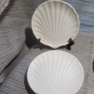 May include: Two white ceramic plates shaped like scallop shells. One plate is positioned upright on a dark brown display stand, while the second plate lies flat in front. Both plates feature a radial ribbed pattern radiating from the base.