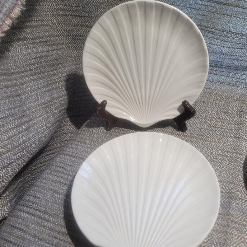 May include: Two white ceramic plates shaped like scallop shells. One plate is positioned upright on a dark brown display stand, while the second plate lies flat in front. Both plates feature a radial ribbed pattern radiating from the base.