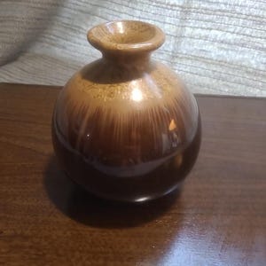 Könnte beinhalten: Eine kleine, kugelförmige Keramikvase mit engem Hals und ausgestelltem Rand. Die Vase hat eine zweifarbige Glasur mit einem glänzenden dunkelbraunen Finish in der unteren Hälfte, das in ein helleres, strukturiertes beige-braunes Tropfmuster übergeht.