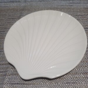 May include: A cream-colored ceramic dish shaped like a scallop shell. The surface features raised, radiating ridges that extend from the base to the curved outer edge, creating a textured, fan-like appearance with a smooth, glossy finish.