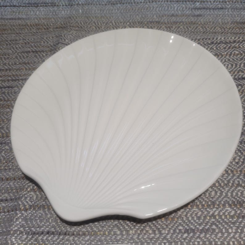 May include: A cream-colored ceramic dish shaped like a scallop shell. The surface features raised, radiating ridges that extend from the base to the curved outer edge, creating a textured, fan-like appearance with a smooth, glossy finish.