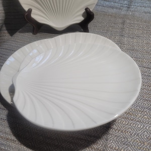 May include: A set of white ceramic plates shaped like seashells, featuring a smooth, glossy finish and raised radial ridges that mimic the natural texture of a scallop shell. The plates are arranged in a stack, with one plate displayed upright.