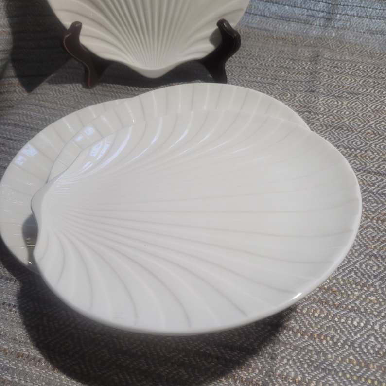 May include: A set of white ceramic plates shaped like seashells, featuring a smooth, glossy finish and raised radial ridges that mimic the natural texture of a scallop shell. The plates are arranged in a stack, with one plate displayed upright.