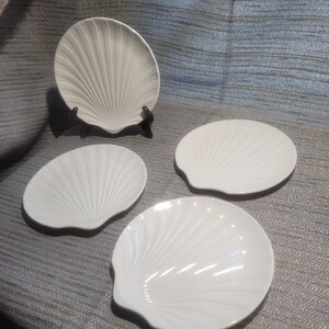 May include: A set of four white ceramic plates shaped like scallop shells. One plate is displayed upright on a small black stand, while the other three plates are arranged flat. Each plate features a radial ribbed pattern mimicking a shell.