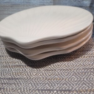 May include: A stack of four off-white ceramic plates shaped like seashells. Each plate features a smooth, glossy finish with raised, radiating ridges that mimic the natural texture and fan-like structure of a scallop shell, creating a uniform set.