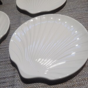 May include: A set of white ceramic plates shaped like scallop shells. Each plate features a glossy finish and raised, radiating ridges that mimic the natural texture of a seashell, extending from the base to the scalloped outer edge.
